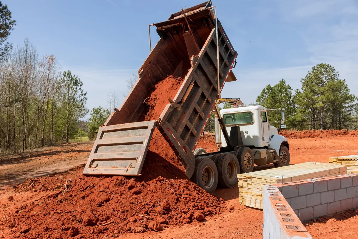 Dump truck unloading a load of red clay fill beside a block foundation